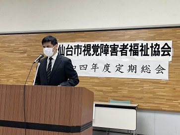 マイクを前に挨拶する高橋会長の後ろに仙台市視覚障害者福祉協会定期総会の横断幕