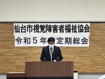 マイクを前に挨拶する高橋会長の後ろに仙台市視覚障害者福祉協会令和5年度定期総会の横断幕