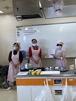 調理台を前に説明するエプロンとマスクをした3人の講師、頭には三角巾