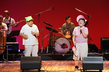 左に合の手を入れる夫の厚さん、右に歌う利枝子さん、後ろにバンド演奏者