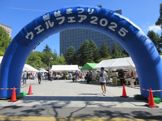 �����܂�E�G���t�F�A2025�Ə����ꂽ�A�[�`�`�̊Ŕ̌��������Ƀe���g�𒣂����e�u�[�X�A�w�i�Ɍ����̌�����������